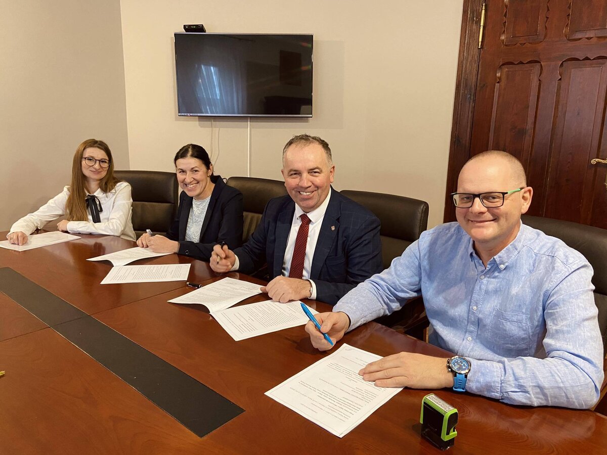 zdjęcie przedstawia cztery osoby siedzące przy stole i podpisujące umowę